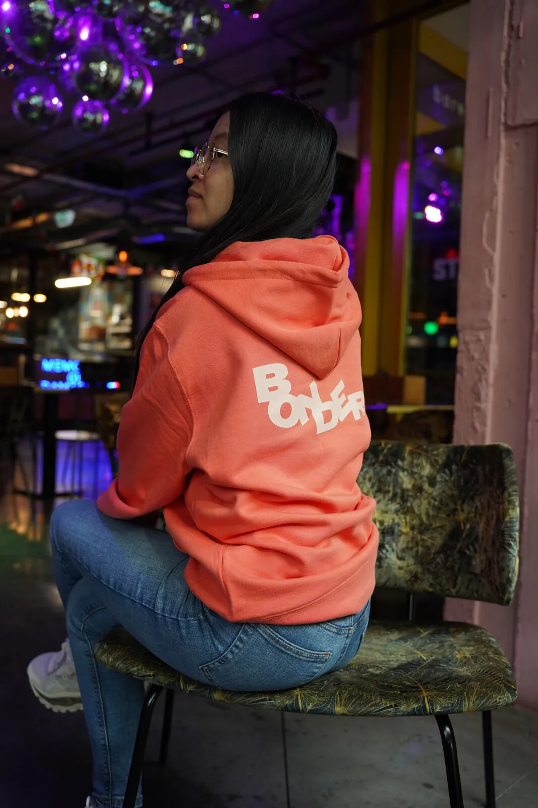 De koraalrode hoodie van opzij, op een stoel voor een verlichte bar