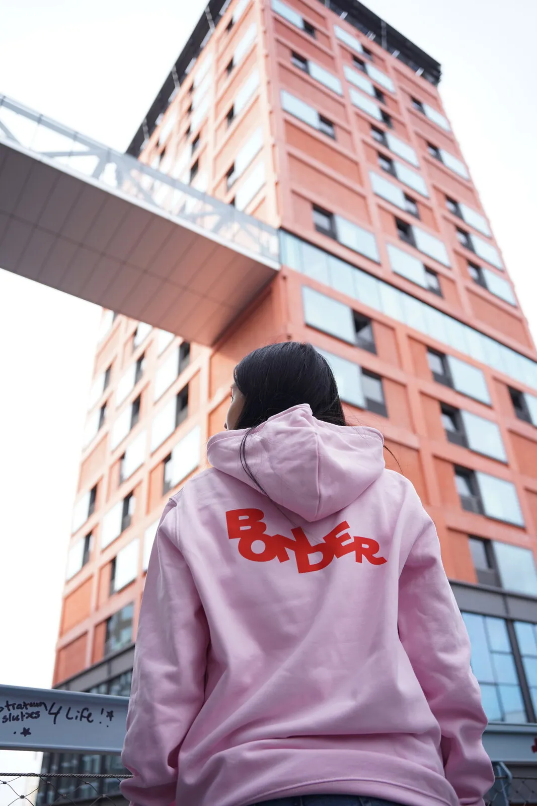 De roze Bonder-hoodie van achteren, met het rode logo op de rug, voor een rode gevel