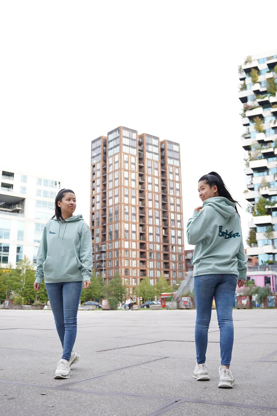 Twee keer de saliegroene hoodie, voor en achter, tussen hoge gebouwen