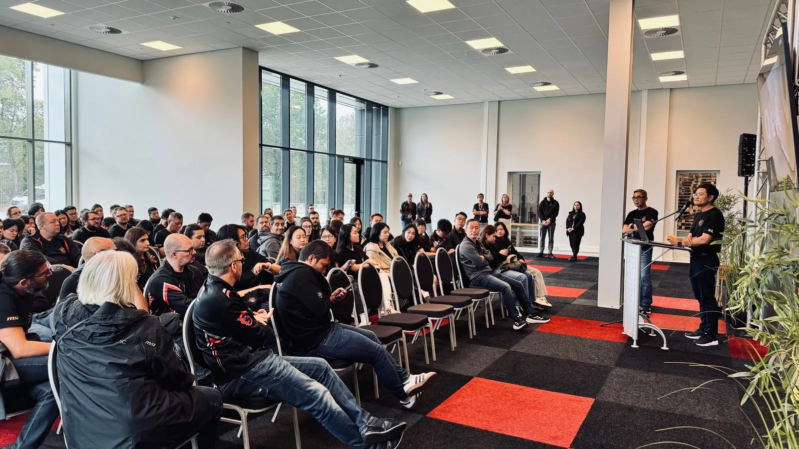 De zaal vol, op een rood en zwart geblokte vloer, luisterend naar de spreker