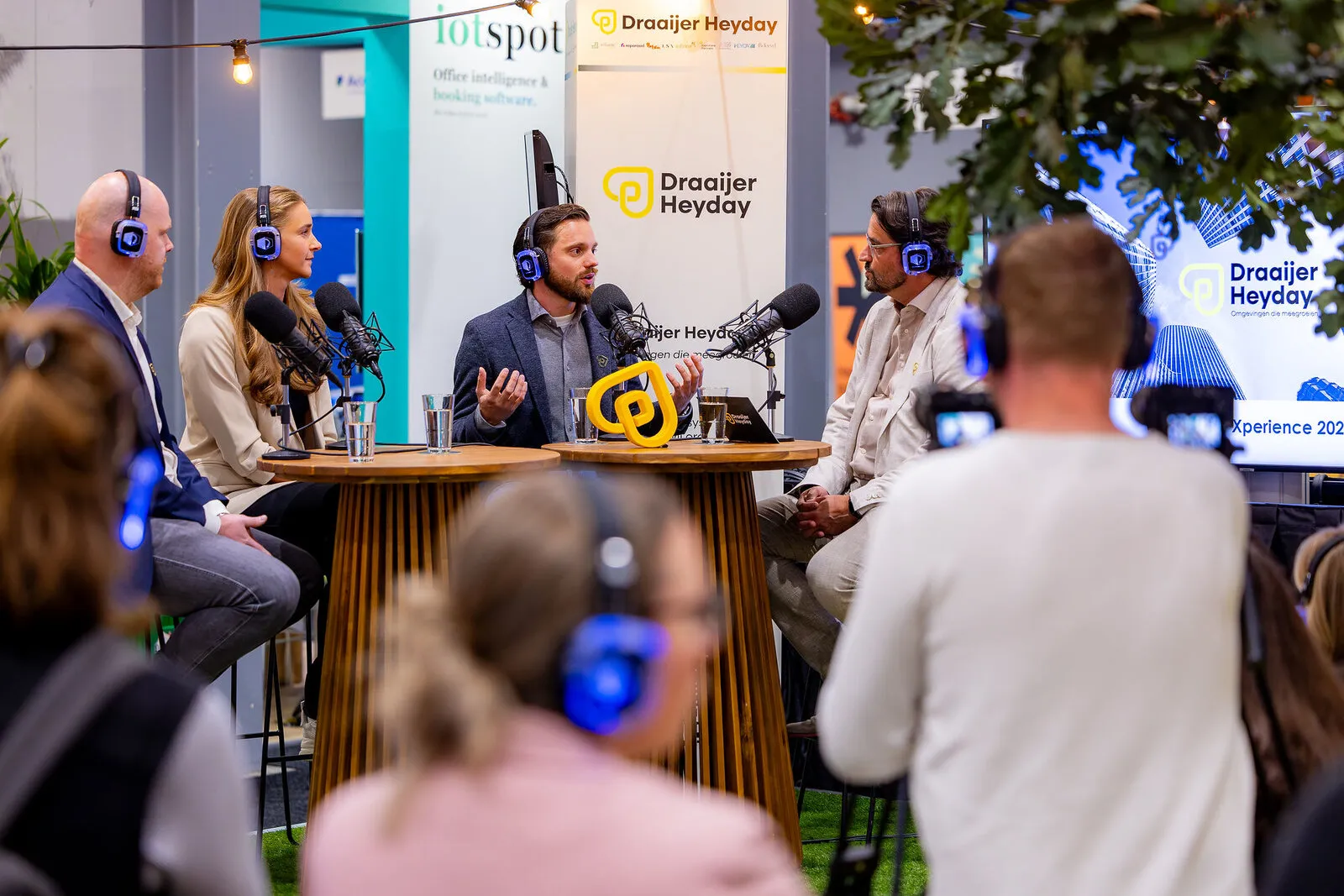 Een live podcastopname op de stand van Draaijer Heyday, met het gele beeldmerk op tafel en publiek met koptelefoons op de voorgrond