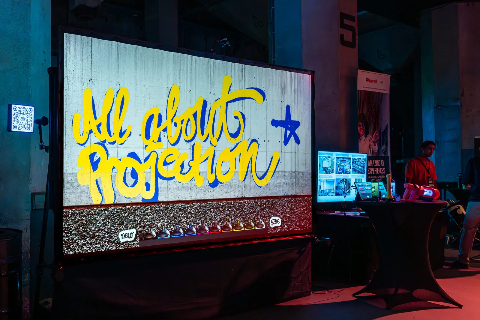 All About Projection sprayed in light across a projected concrete wall, a rack of spray cans below it